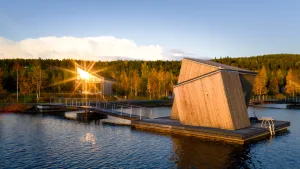 Angular floating cabin on the Lule River at golden hour connected by a wooden walkway to a glass-fronted land building amid autumn-colored forest at Arctic Bath in Swedish Lapland