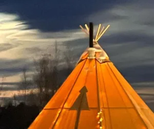 Glowing orange Sámi lavvu tent at dusk with wooden poles and chimney against a cloudy sky